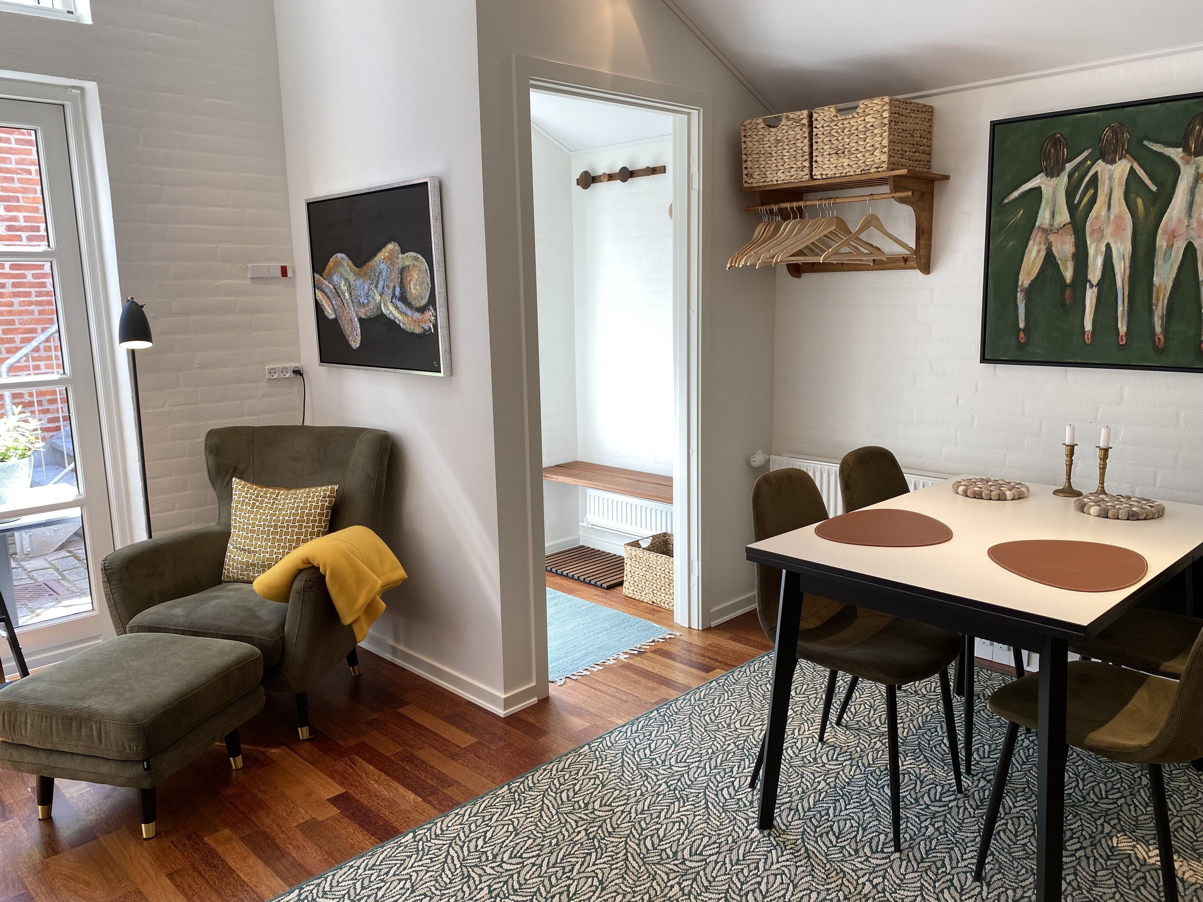 A modern living room with a grey armchair, ottoman, and a dining table with chairs. There is a painting on the wall and a basket on the shelf.