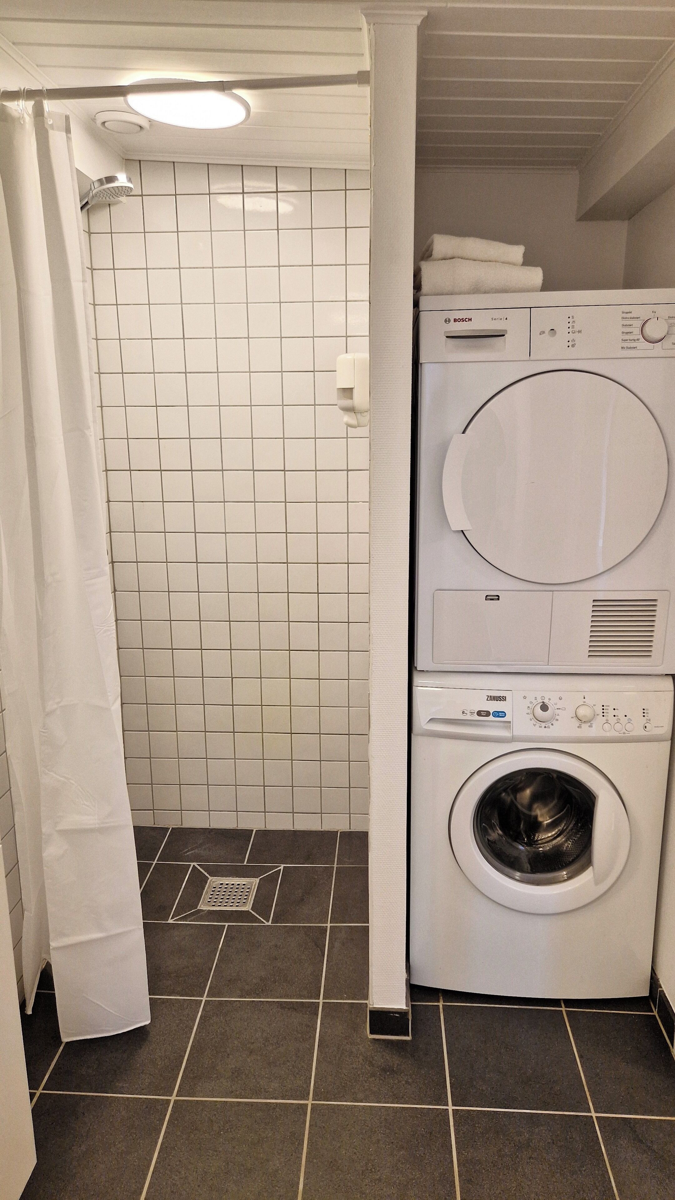 A compact laundry area with a Bosch washing machine and dryer, a tiled shower area, and a drain grate on the floor.