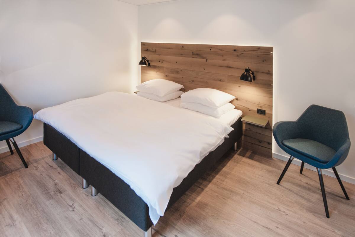 A modern bedroom with a wooden headboard, a large bed with white linens, two bedside tables with lamps, and two blue chairs.