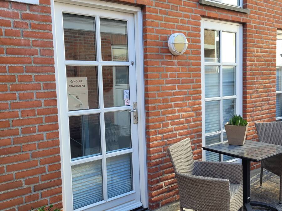 Een gebouw van baksteen met een raam met witte omlijsting en een deur, een klein buitentafeltje met rieten stoelen en een bloempot met plant.