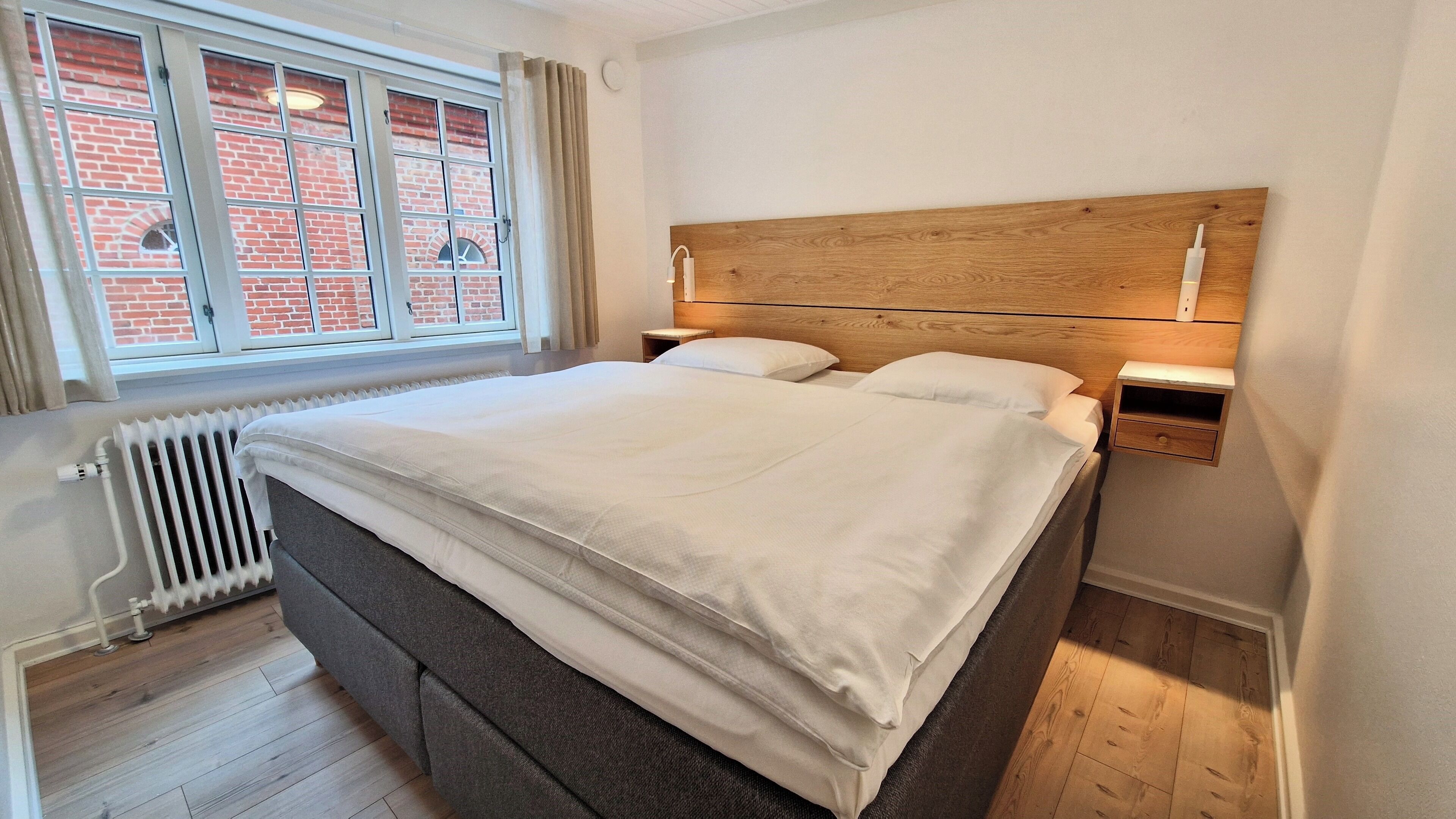 Una cama bien tendida en una habitación con cabecera de madera, una ventana con cortinas, un radiador y una mesita de noche con una lámpara.