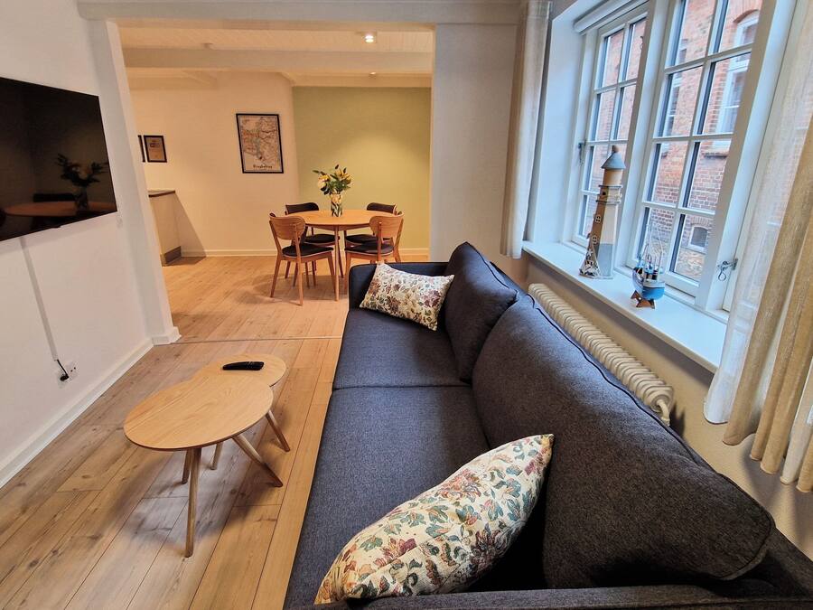 Un salón con un sofá gris oscuro, dos cojines estampados, una pequeña mesa de madera y un televisor montado en la pared.