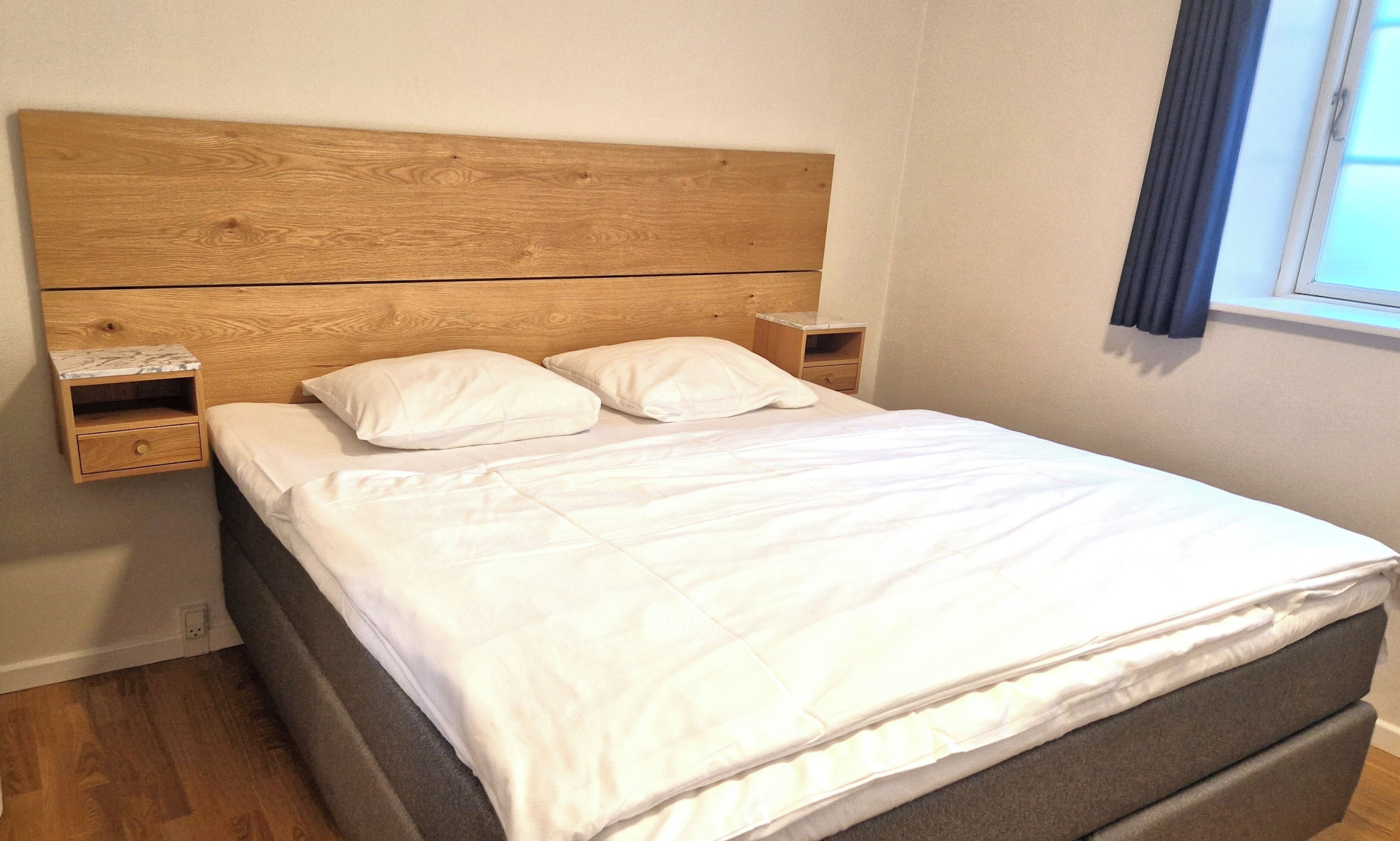 A neatly made bed with a wooden headboard, two bedside tables with drawers, and a window with curtains.