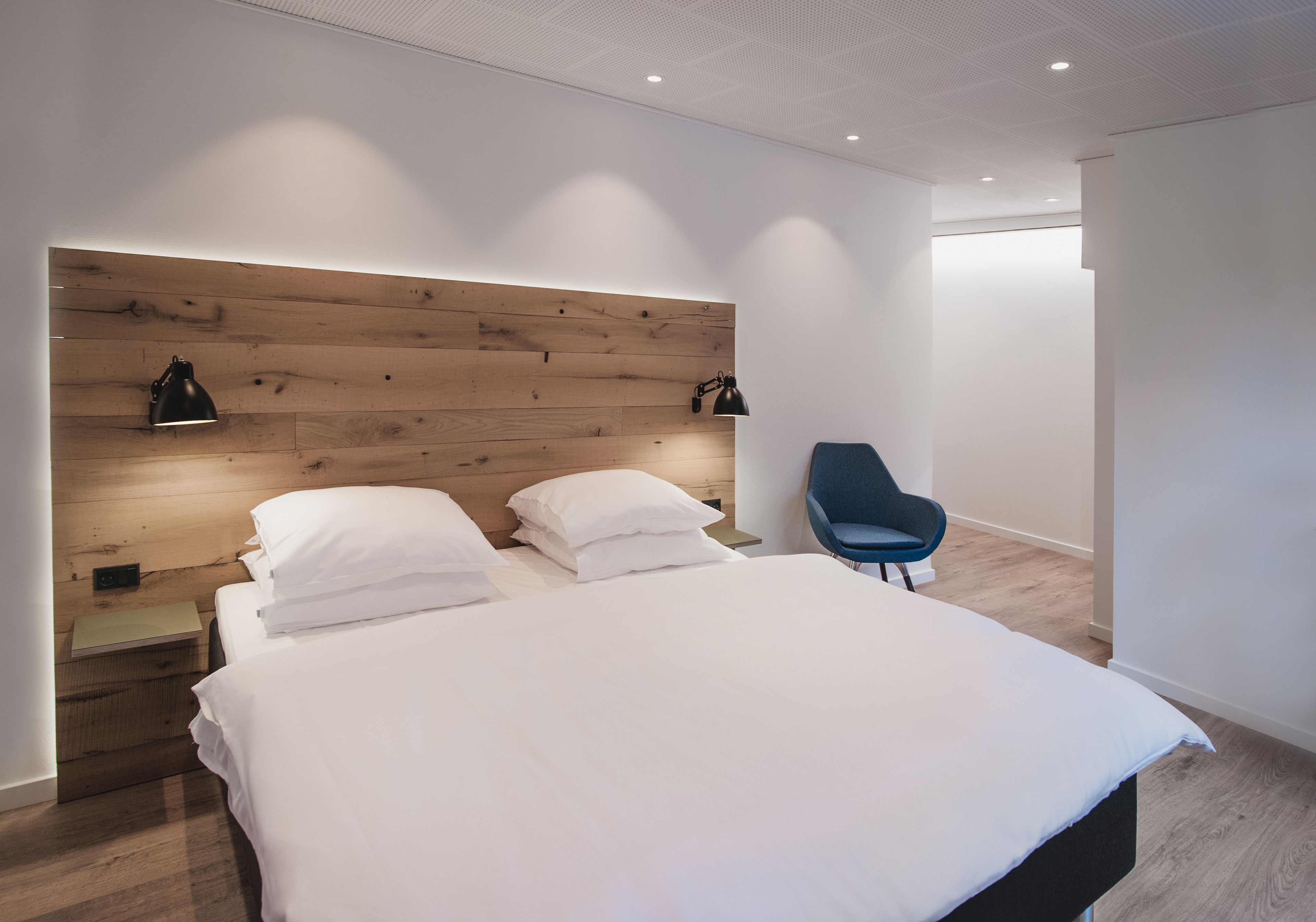 Une chambre moderne avec une tête de lit en bois, un lit simple avec une literie blanche, une chaise bleue et deux lampes fixées au mur.