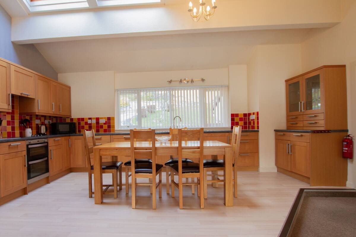 A spacious kitchen with wooden cabinets, a dining table with chairs, a large window with blinds, and a chandelier.