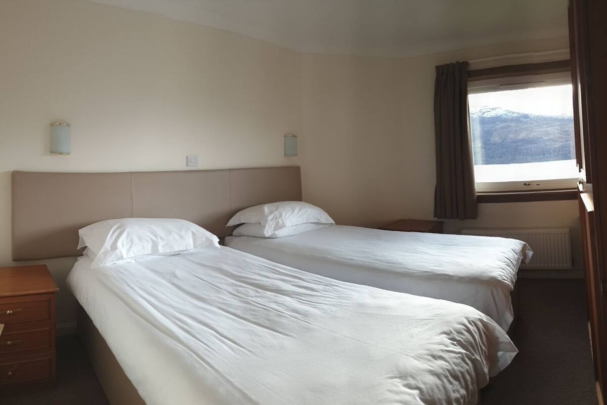 A hotel room with two beds, a wooden nightstand, a window with curtains, and a view of the mountains.