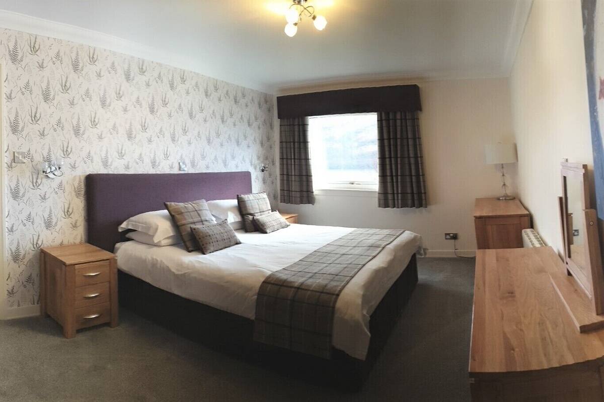 A bedroom with a bed, bedside tables, a window with curtains, a wall-mounted lamp, and a painting.