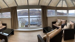 A cozy seating area with wicker furniture and a large window overlooking a lake and mountains. - Loch Rannoch Highland Club (Pitlochry)