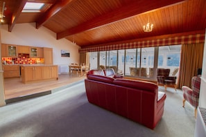 A spacious living area with a red leather sofa, wooden flooring, a kitchen with wooden cabinets, and a large window with a view. - Loch Rannoch Highland Club (Pitlochry)
