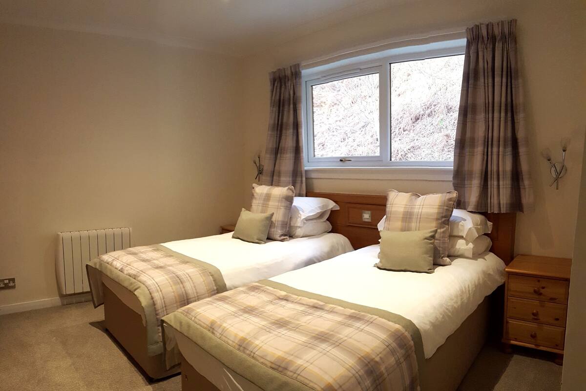 A bedroom with two beds, a window with curtains, a radiator, and a wooden nightstand with drawers.