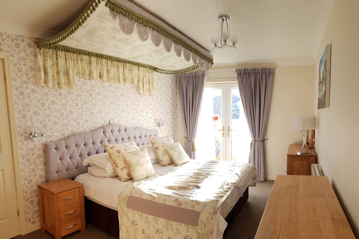 A bedroom with a tufted headboard, floral-patterned bedding, a wooden nightstand, a wooden desk, and a chandelier.