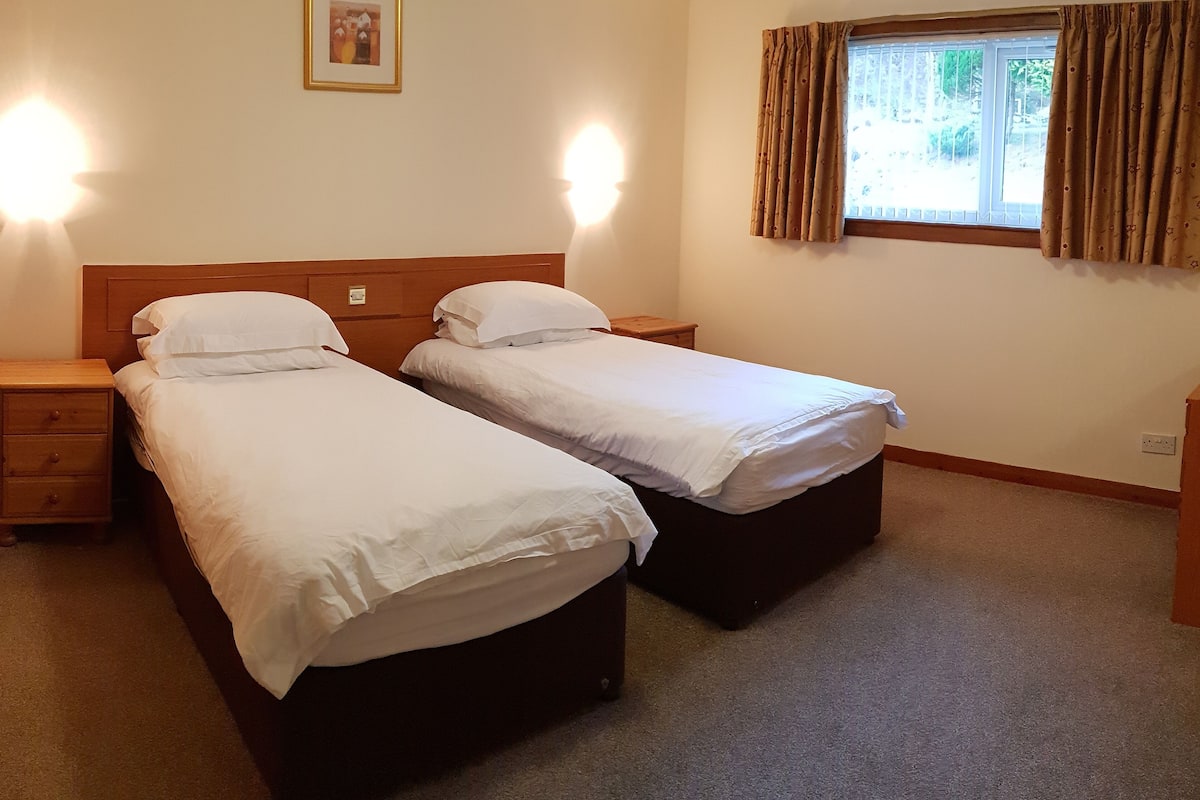 A room with two single beds, a wooden nightstand, a small drawer unit, and a window with curtains.