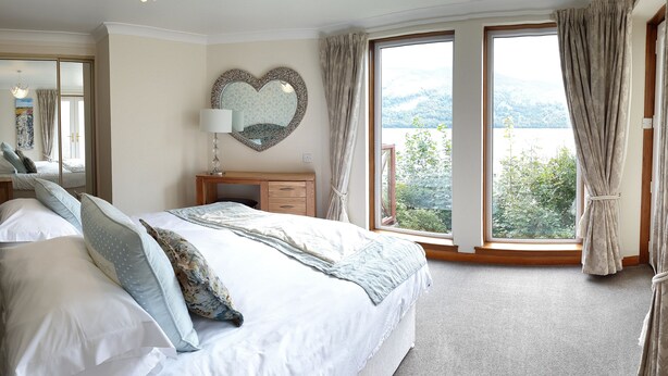 A bedroom with a large bed, a dresser, a mirror, and a view of a lake through large windows. - Loch Rannoch Highland Club (Pitlochry)