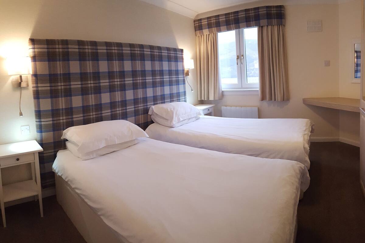 A hotel room with two beds, a nightstand, a lamp, a window with curtains, and a wooden wardrobe.