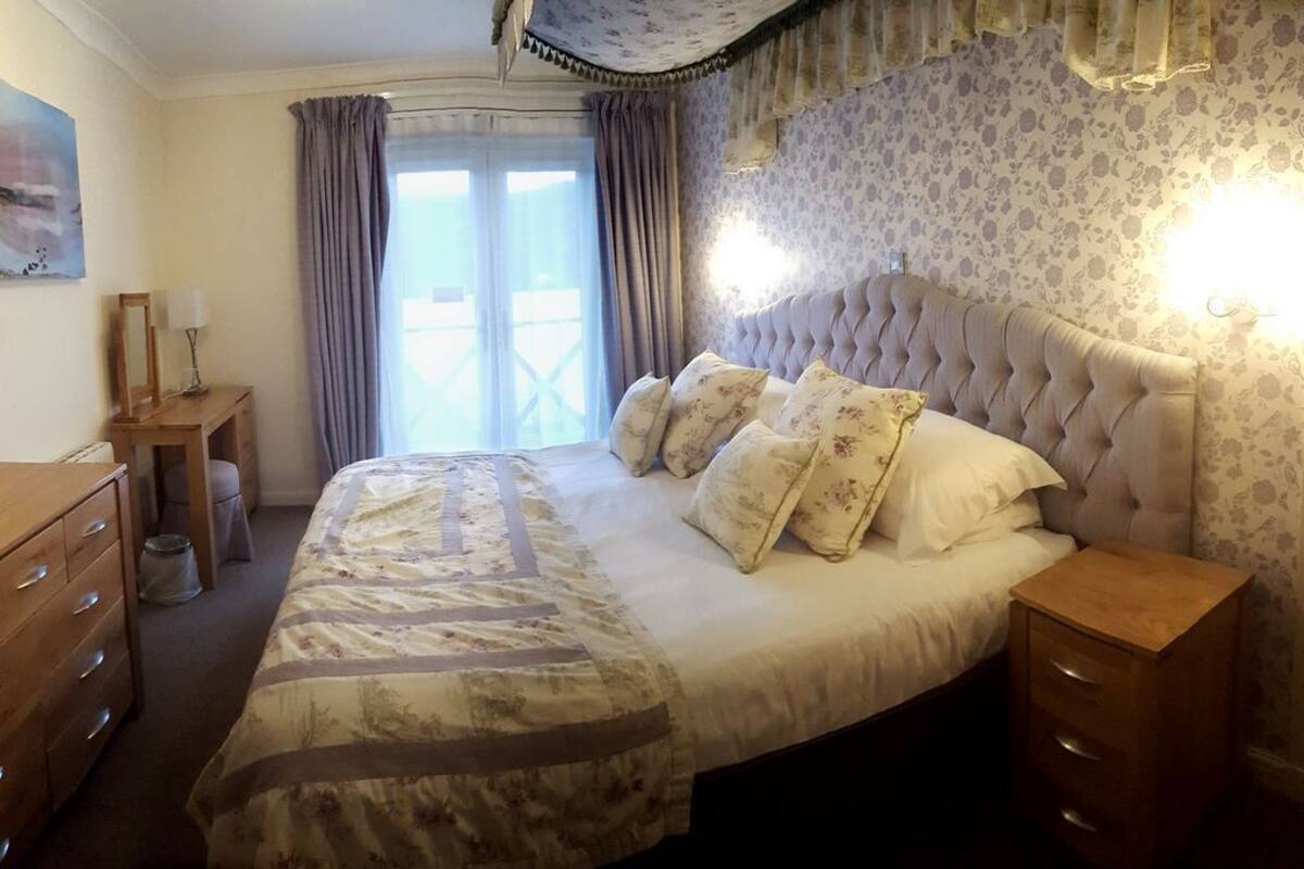 A bedroom with a large bed, a wooden dresser, a nightstand, and a window with curtains.
