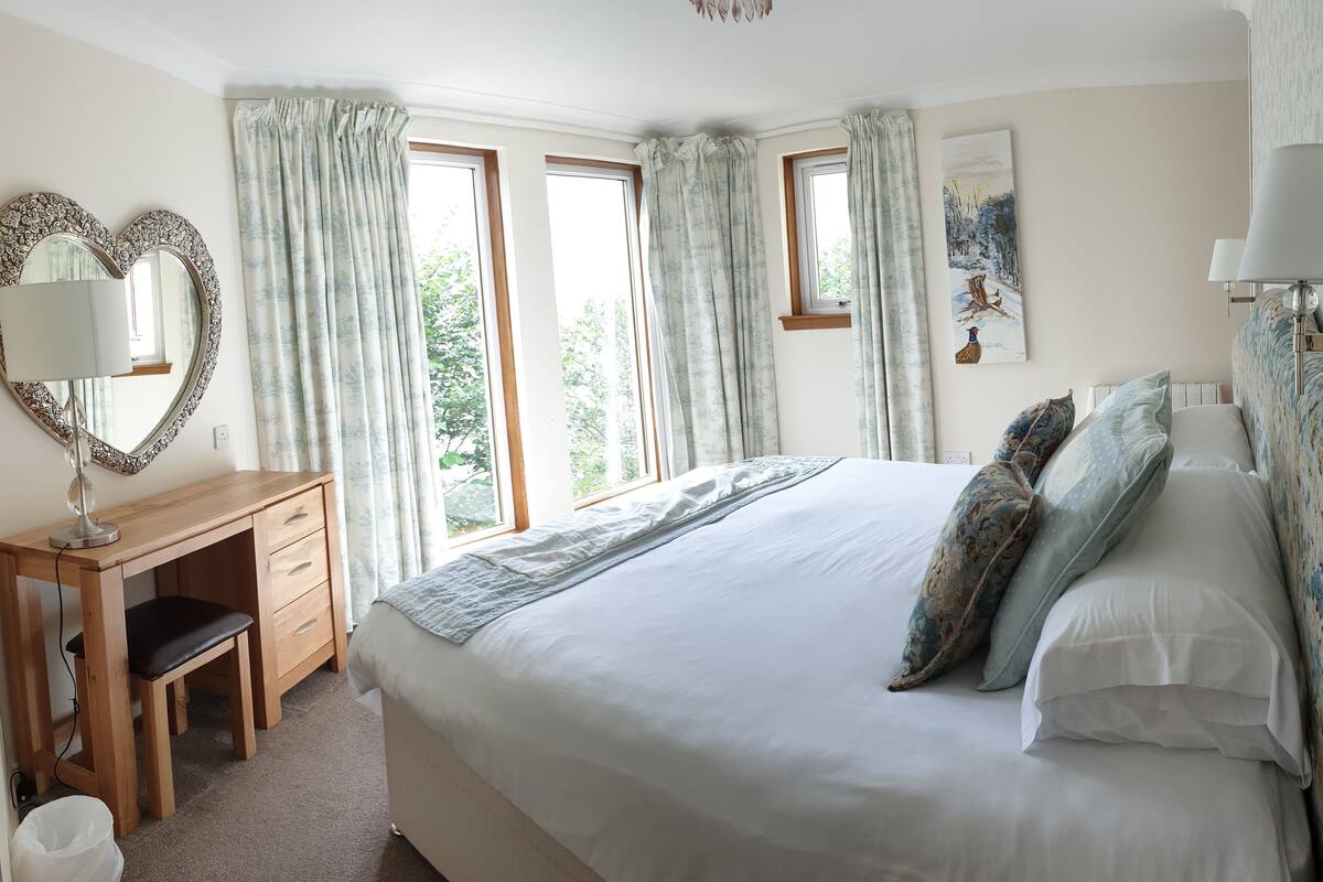 A bedroom with a large bed, a wooden desk, a mirror, and two windows with curtains.