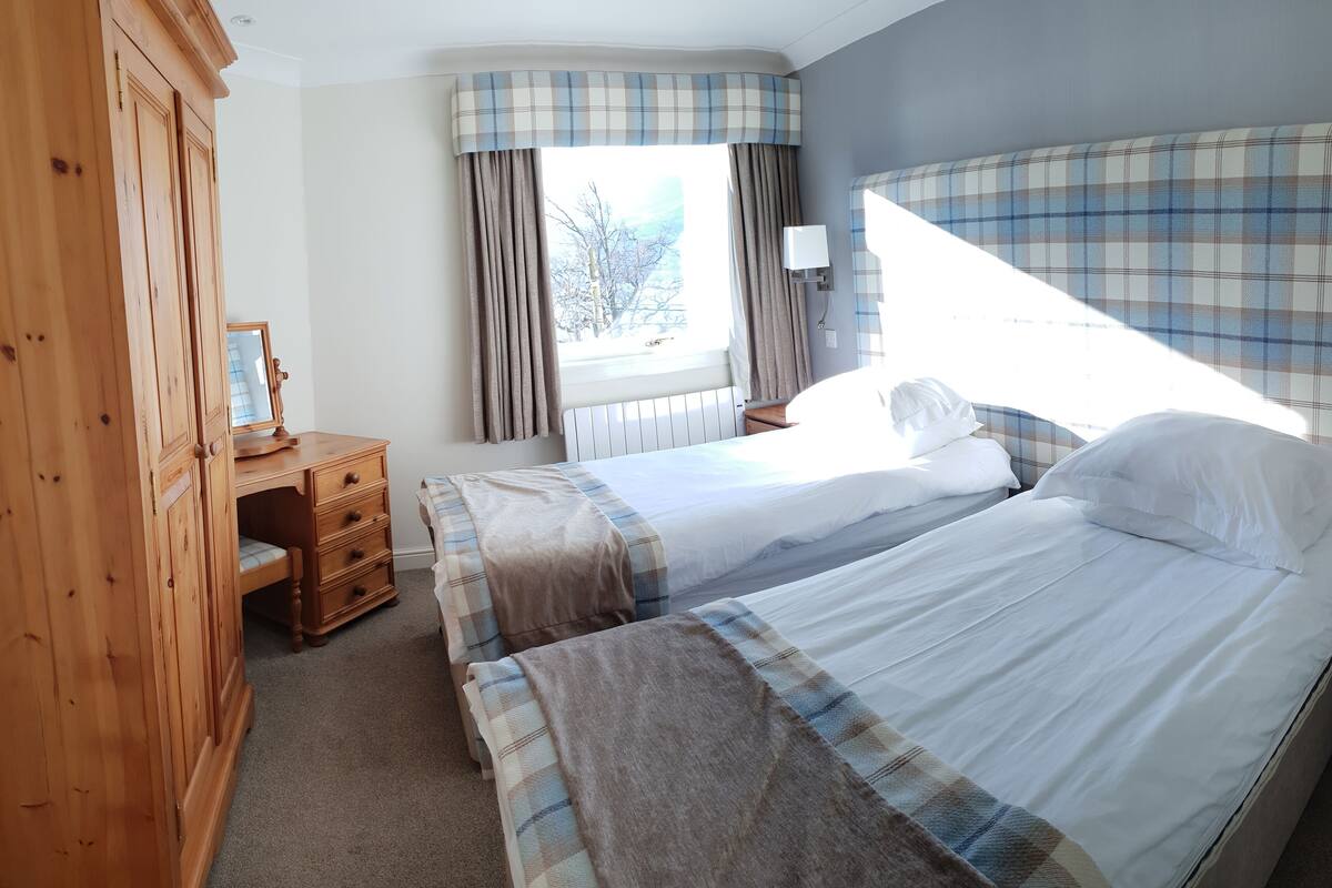 A bedroom with two beds, a wooden wardrobe, a bedside table with a lamp, and a window with curtains.