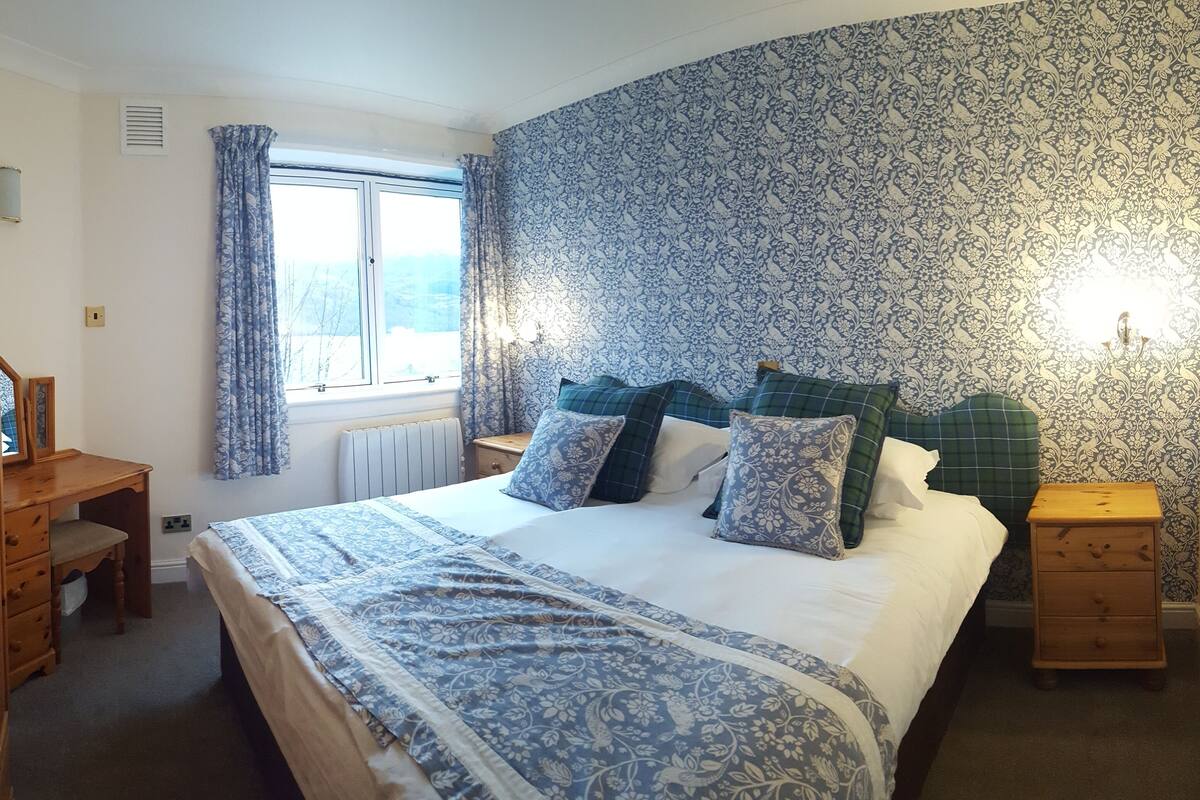 A bedroom with a large bed, a wooden dresser, a nightstand, and a window with curtains.
