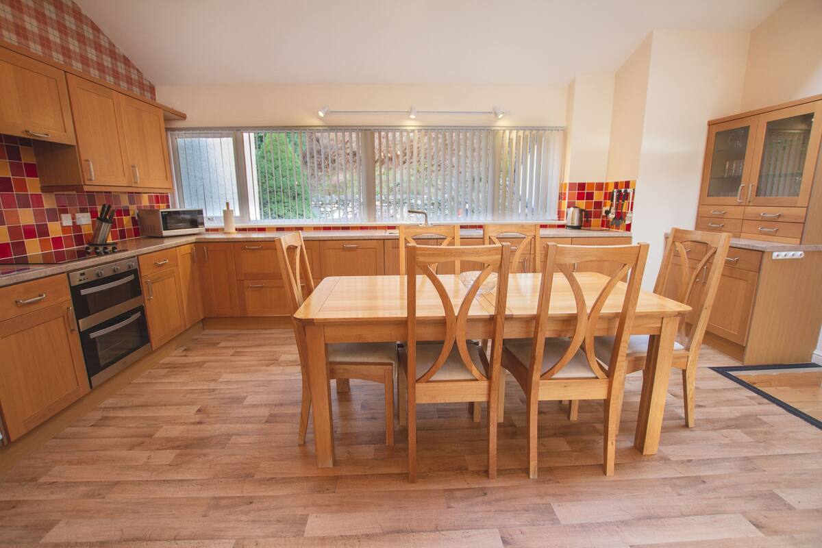 A kitchen with wooden cabinets, a dining table with chairs, a microwave, and a window with blinds.