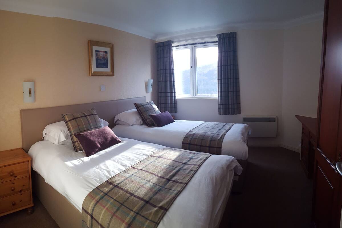 A hotel room with two beds, a nightstand, a window with curtains, and a wall-mounted light switch.