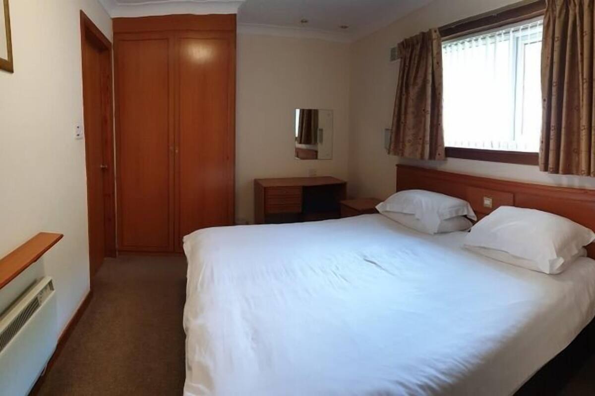 A hotel room with a large bed, a wooden wardrobe, a bedside table, a small desk, and a window with curtains.