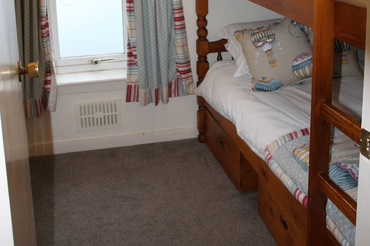 A small, single-bedded room with a window and curtains, a radiator, and a bunk bed with patterned bedding.