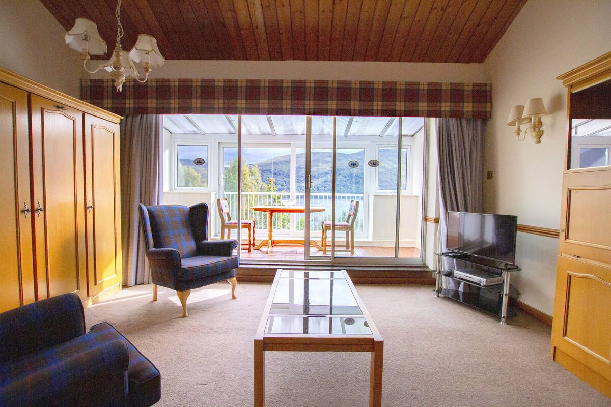 A living room with a glass-top coffee table, plaid armchairs, a sofa, a television on a stand, and a view of a balcony with a table and chairs.