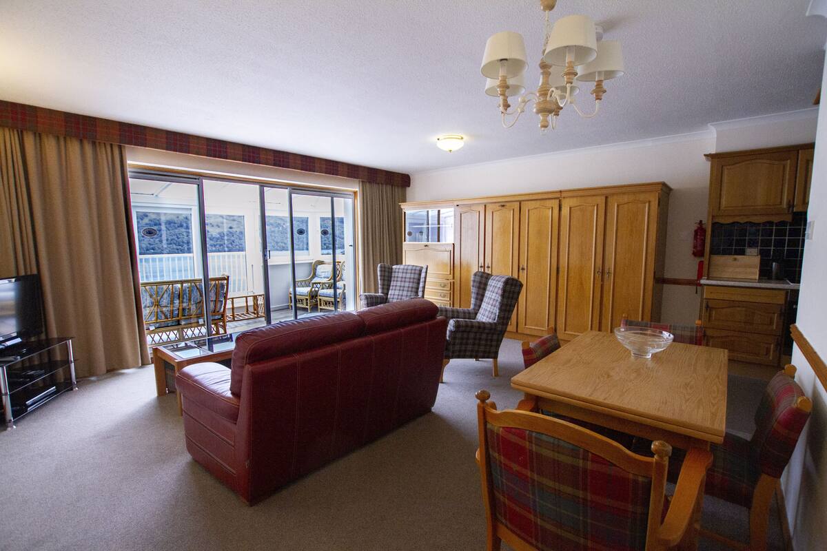 A spacious living area with a red sofa, wooden dining table, and plaid chairs. There is a large sliding glass door leading to a balcony.