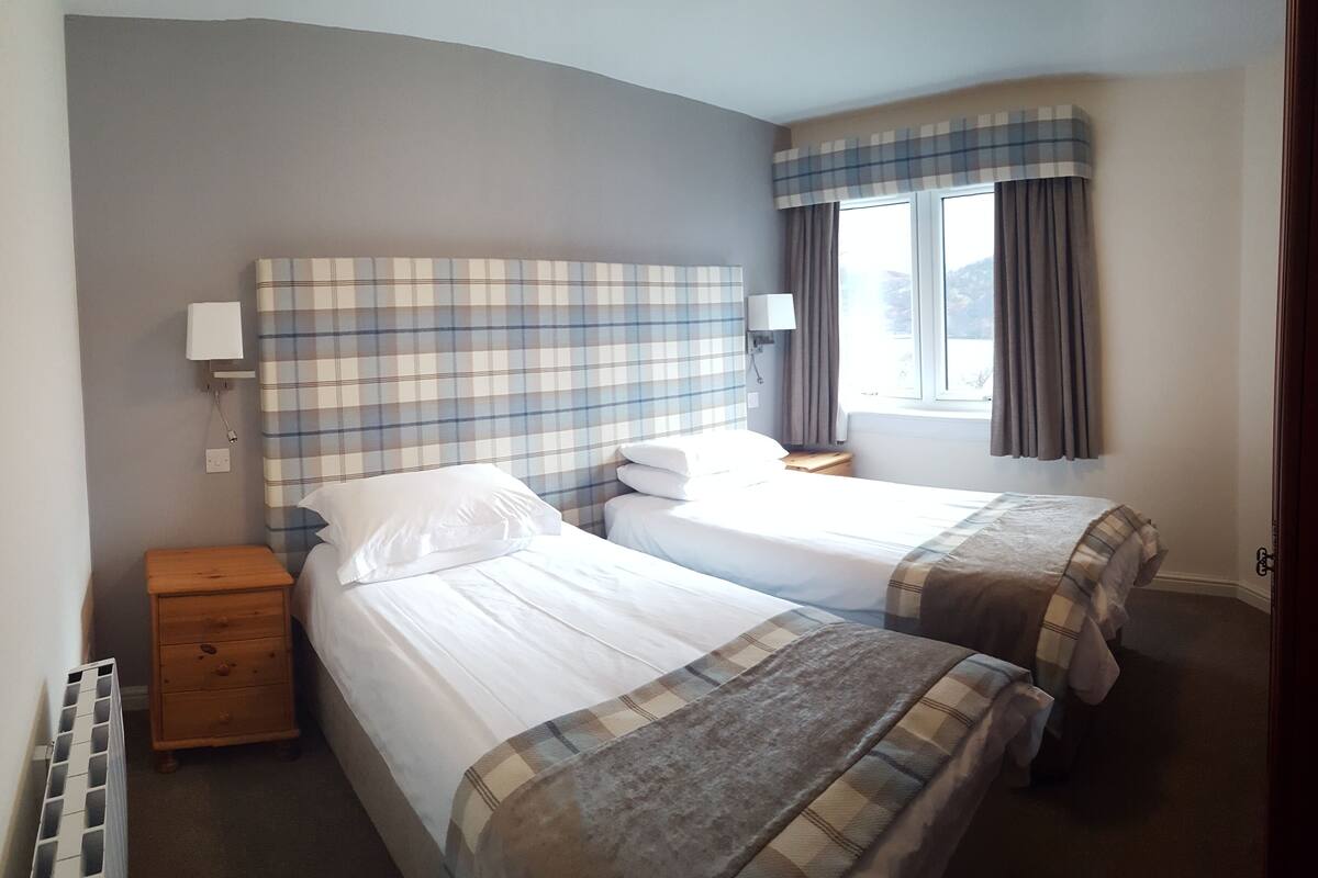 A hotel room with two beds, a nightstand, a lamp, and a window with curtains.