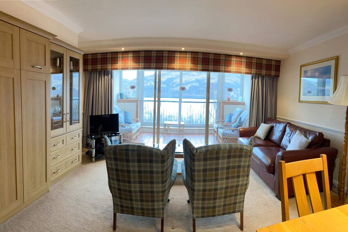 A living room with a sofa, two plaid chairs, a TV, and a balcony with curtains.