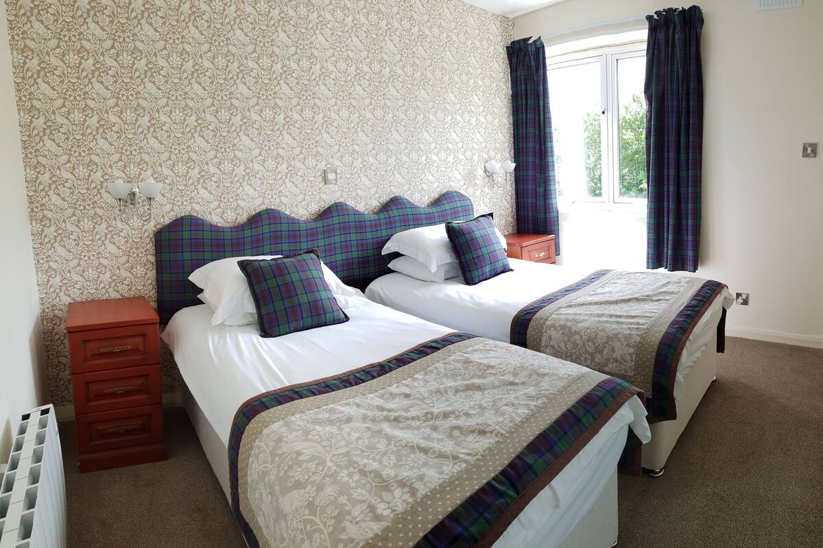 A double room with two beds, each with plaid bedding and headboards, a wooden nightstand, and a radiator.