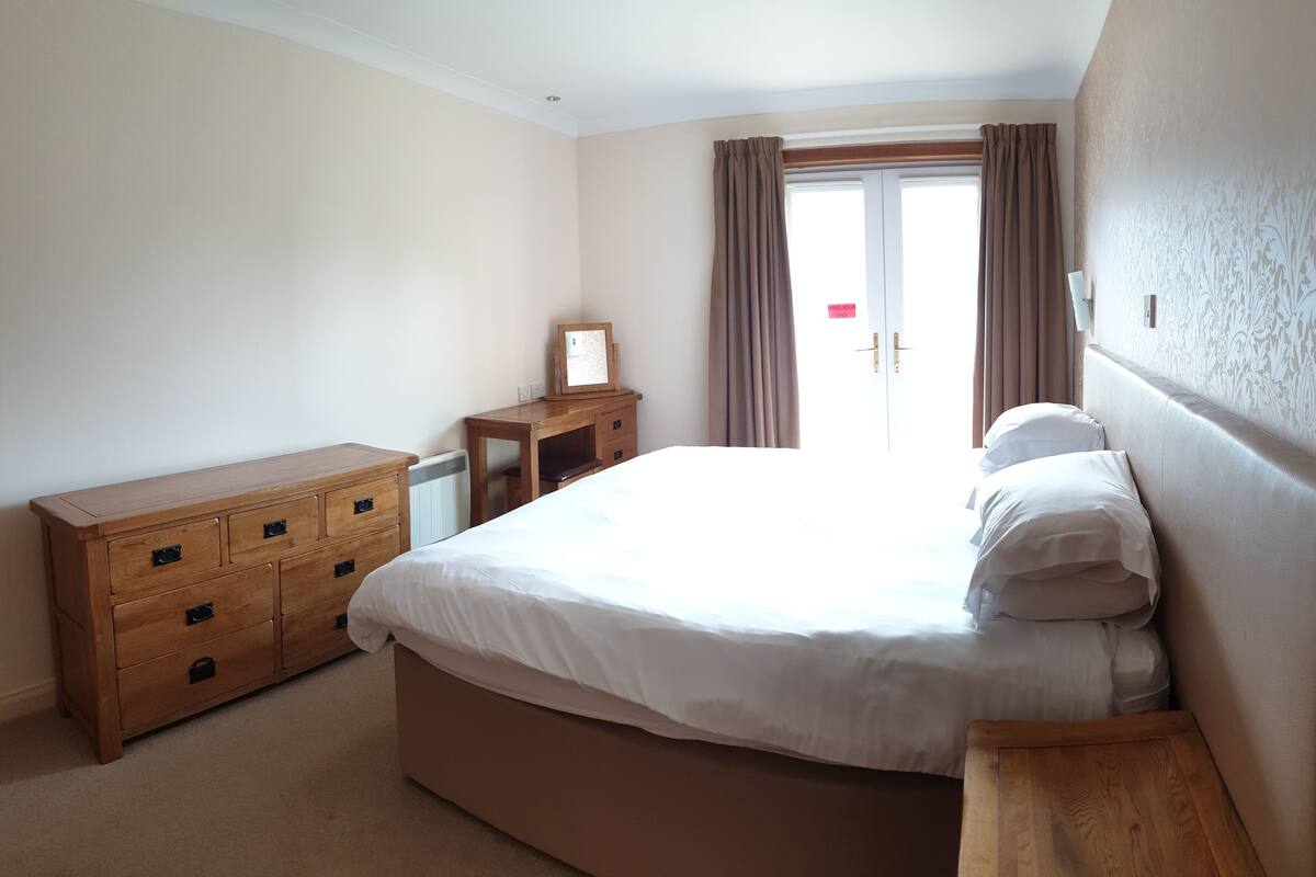 A bedroom with a large bed, a wooden dresser, a desk, and a window with curtains.