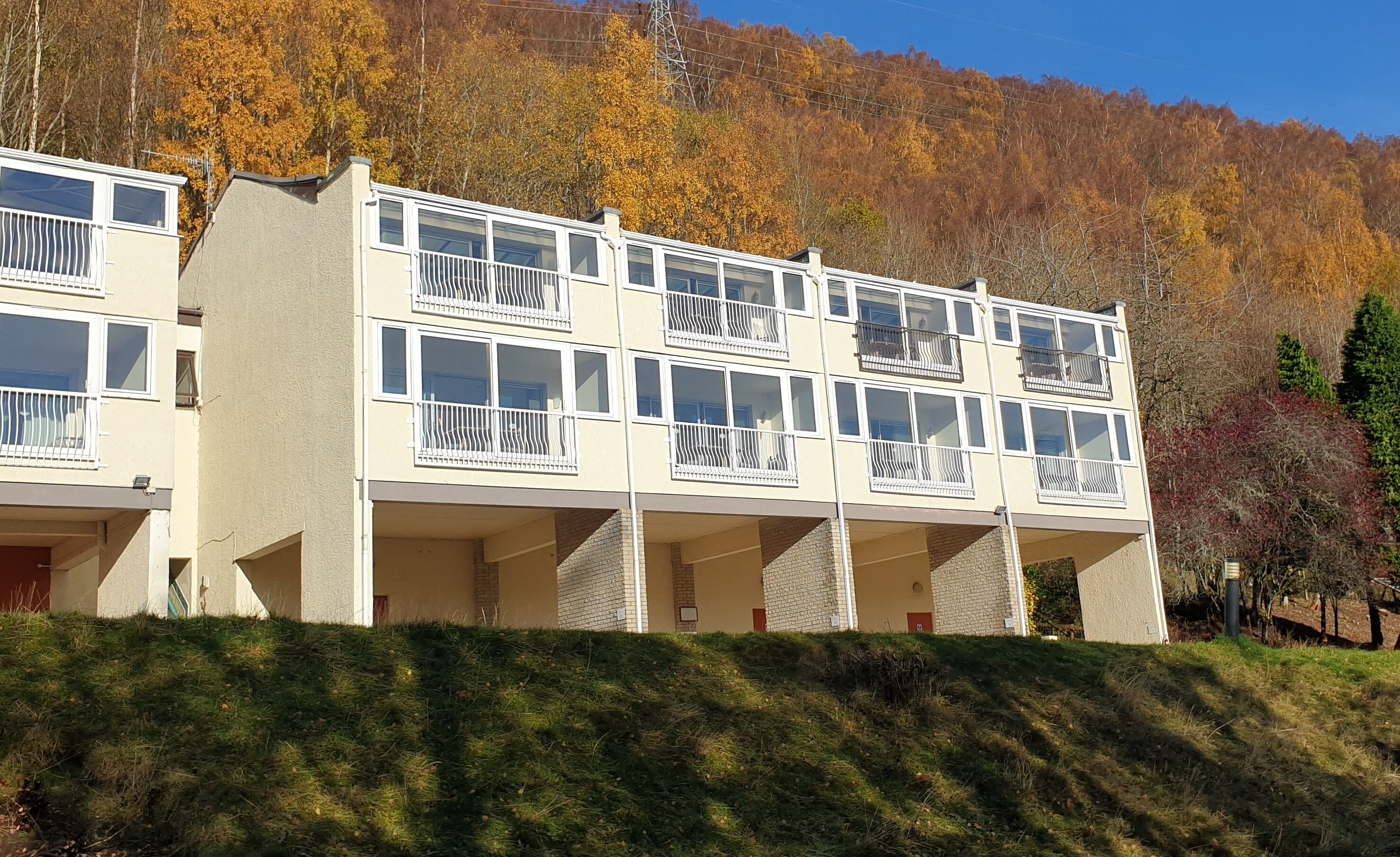 A multi-story building with balconies, set against a backdrop of autumn-colored trees.
