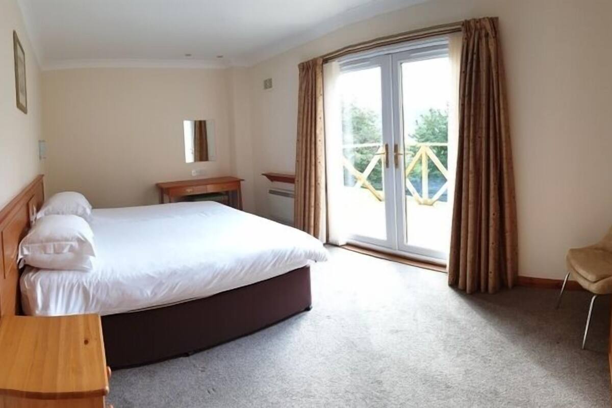A hotel room with a large bed, a wooden nightstand, a small desk, and a chair. There is a balcony with curtains.