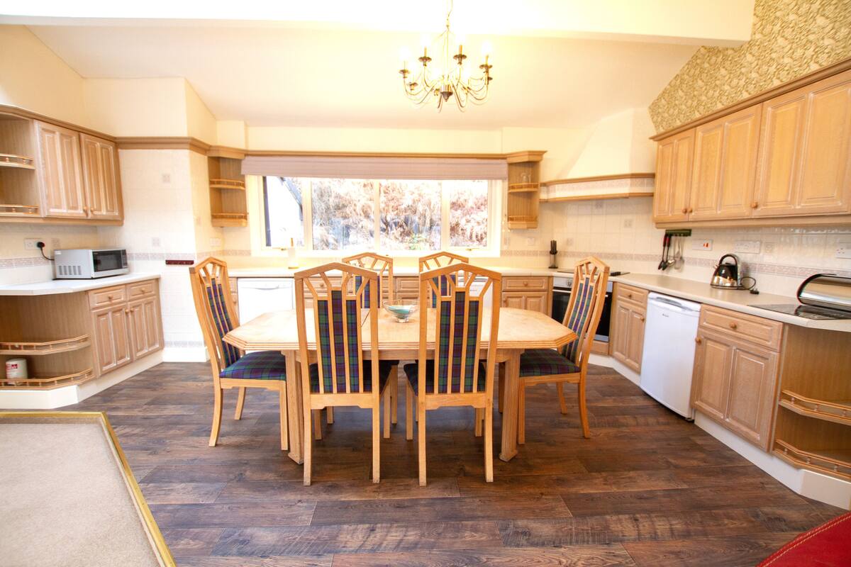 A kitchen with wooden cabinets, a dining table with chairs, a chandelier, and a window with a view of trees.