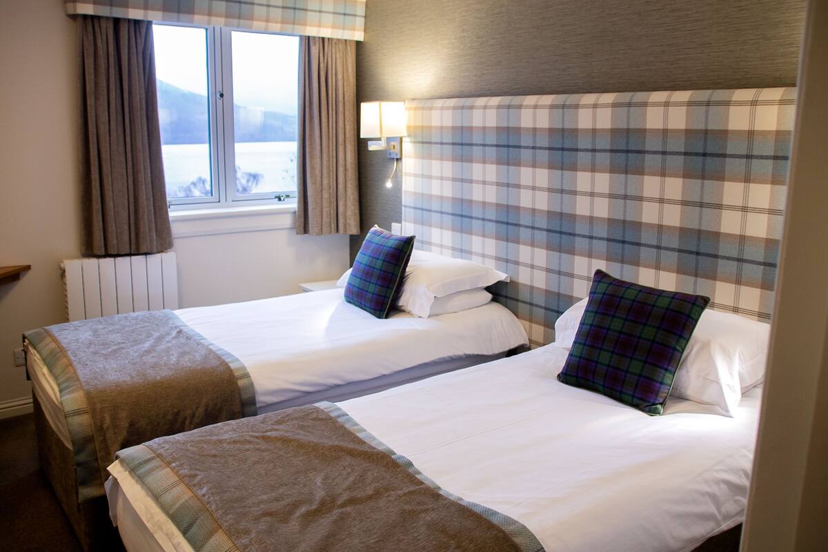 A hotel room with two beds, a window with curtains, a wall-mounted lamp, and a view of the outdoors.
