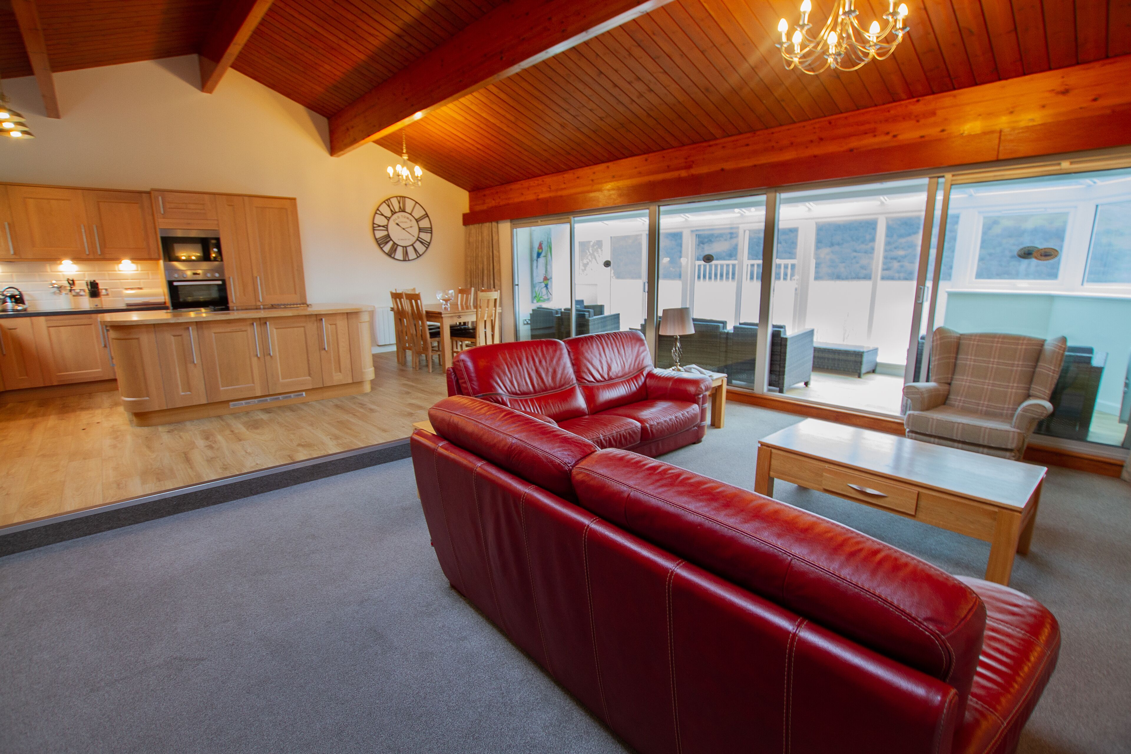 A spacious living area with a red leather sofa, a wooden coffee table, and a dining table with chairs. There is a kitchen in the background with wooden cabinets and a clock on the wall.