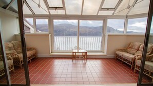 A sunroom with a view of a lake and mountains, furnished with wicker chairs and a small table. - Loch Rannoch Highland Club (Pitlochry)
