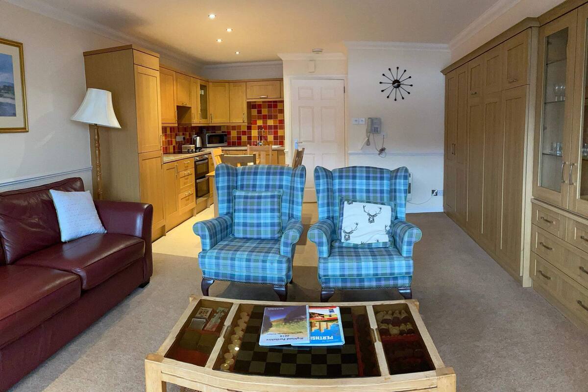 A living room with a red leather sofa, two plaid armchairs, a coffee table with magazines, and a kitchen in the background.