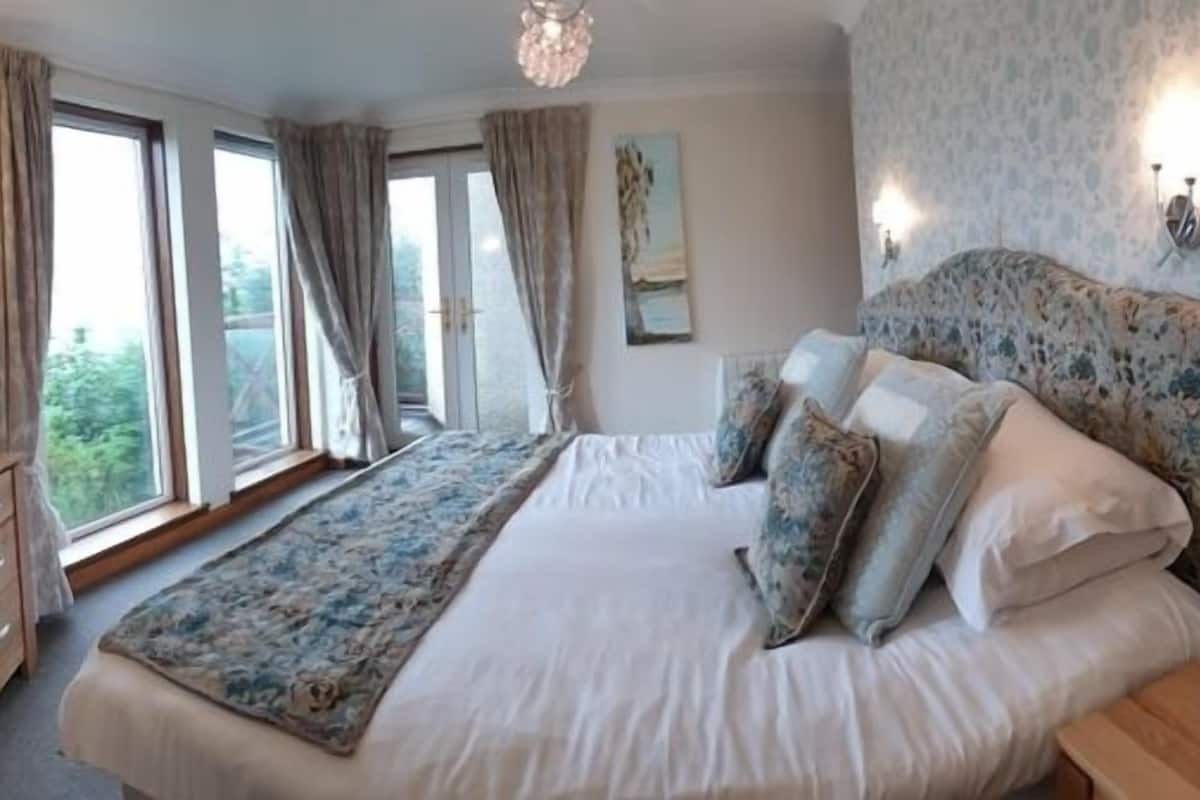 A bedroom with a large bed, a wooden desk, and a view of the outdoors through large windows.