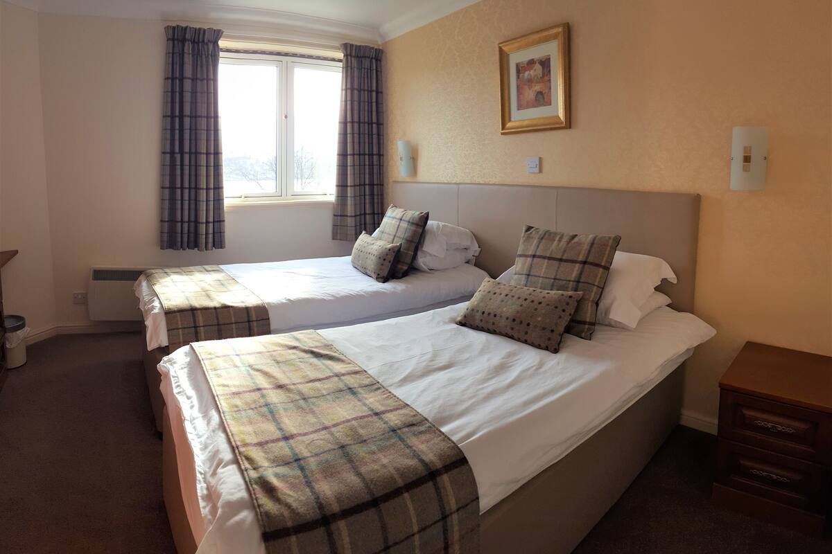 A hotel room with two single beds, a large bed, a window with curtains, a framed picture on the wall, and a small wooden nightstand.