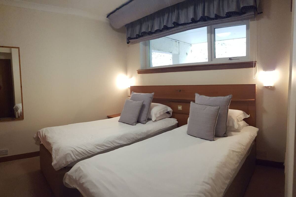 A hotel room with two single beds, a window with curtains, and wall-mounted lights.