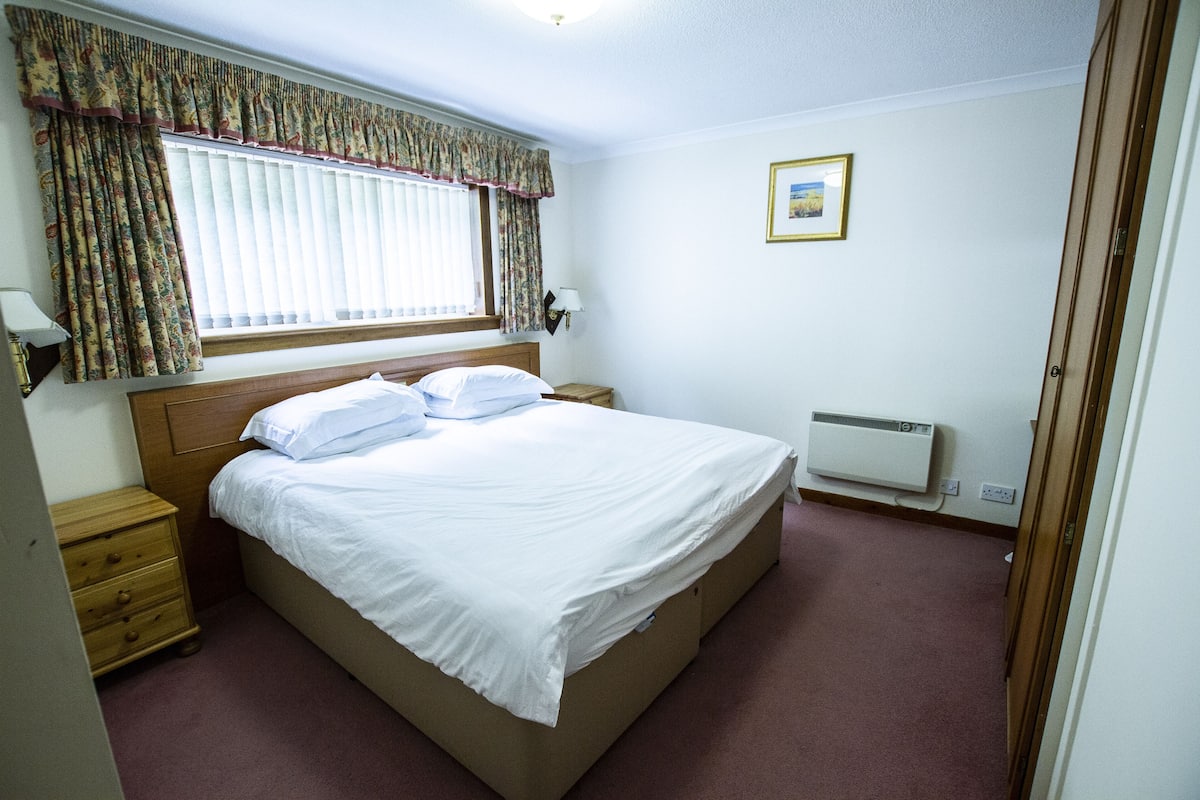 A bedroom with a single bed, a nightstand, a dresser, a window with curtains, a wall-mounted lamp, and a framed picture.
