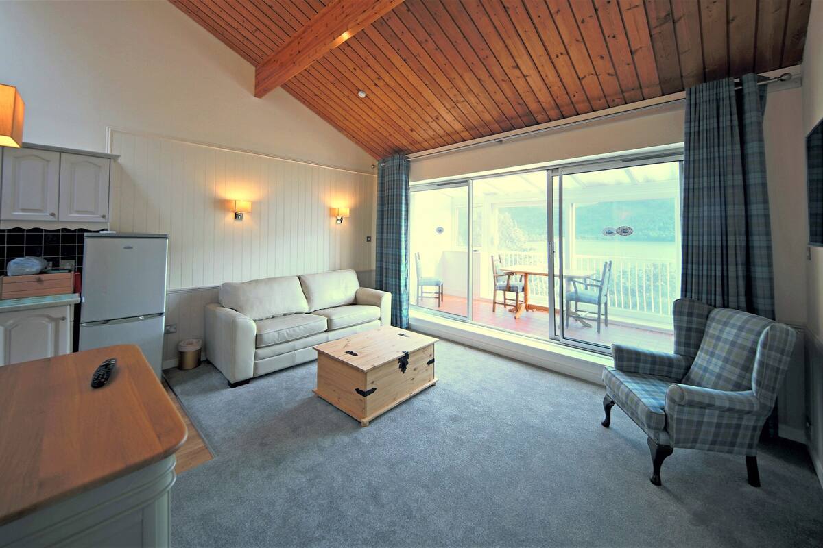 A cozy room with a wooden ceiling, a sofa, a plaid armchair, a small table, and a sliding glass door leading to a balcony with outdoor seating.