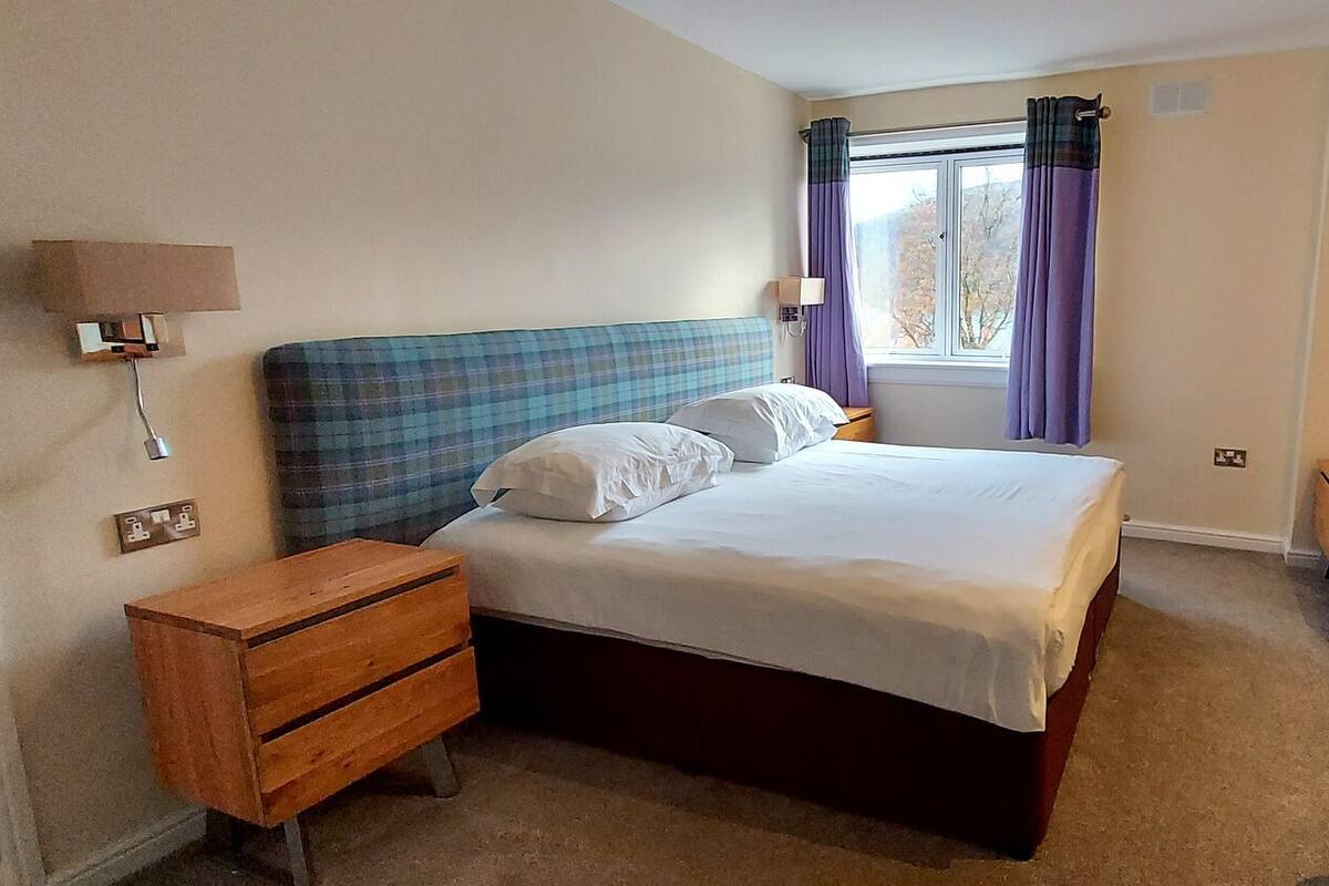 A bedroom with a bed, a wooden nightstand, a desk, and a window with curtains.