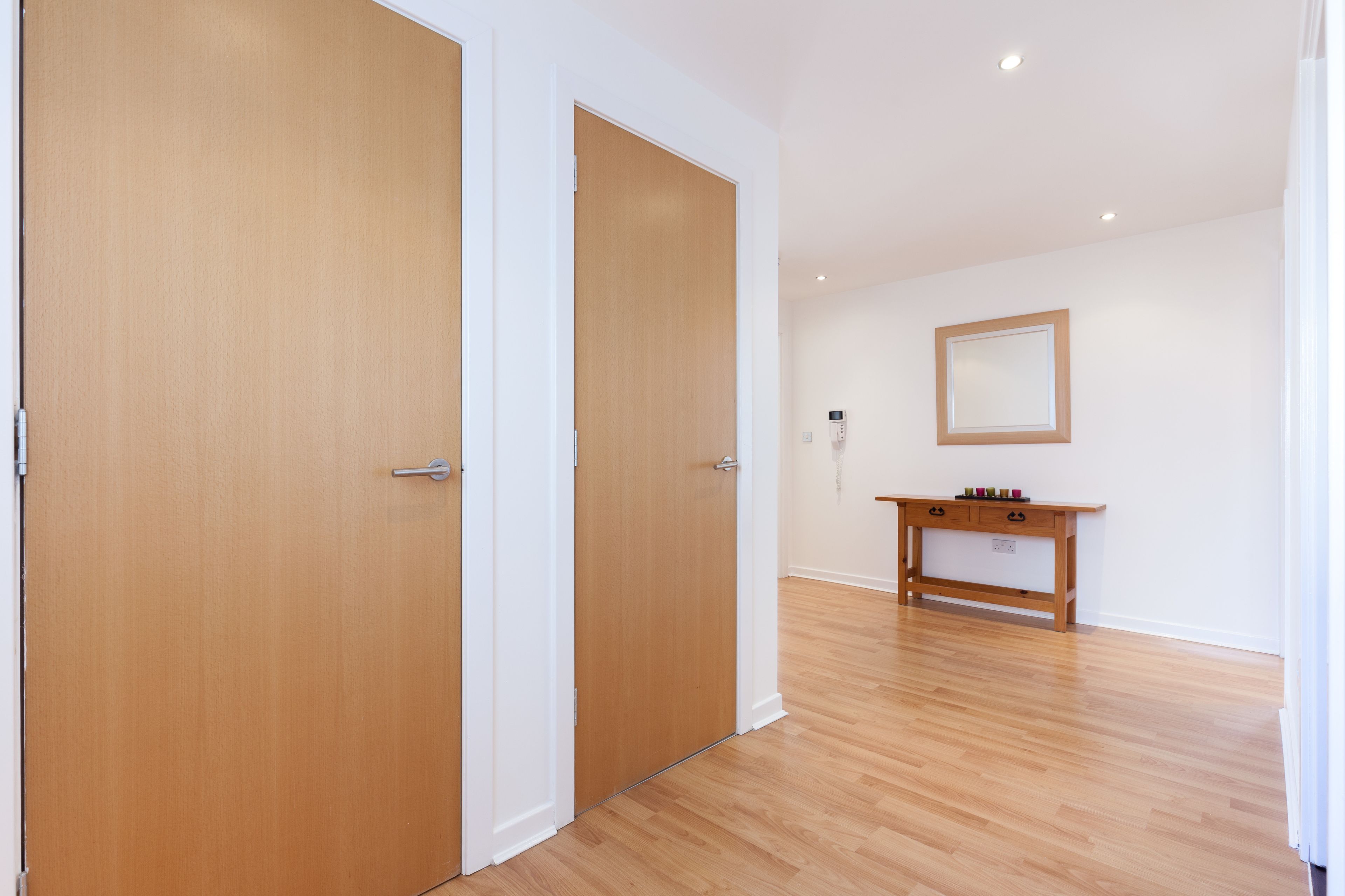 Een kamer met houten vloer, twee gesloten deuren, een klein houten console met lades en een spiegel aan de muur.