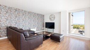 A modern living room with a stone wall, leather sofa, glass coffee table, and a large window offering a view. - Stylish Garden View Apartment (Edinburgh)