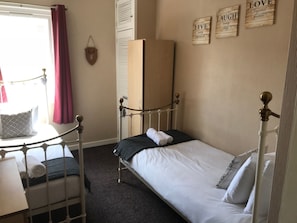 Ein Einzelbett mit weißer Matratze und Kissen, ein Holzschrank, ein kleiner Tisch mit Lampe sowie ein Fenster mit Vorhängen.