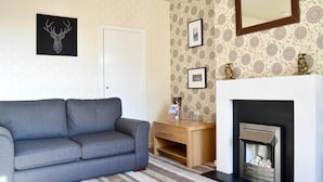 A living room with a grey sofa, a wooden side table, a fireplace, and patterned wallpaper. - Crags View Apartment (Edinburgh)
