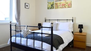 A neatly made bed with a black metal frame, white bedding, and a grey throw blanket. There are two bedside tables with lamps, a window with curtains, and floral artwork on the wall. - Crags View Apartment (Edinburgh)